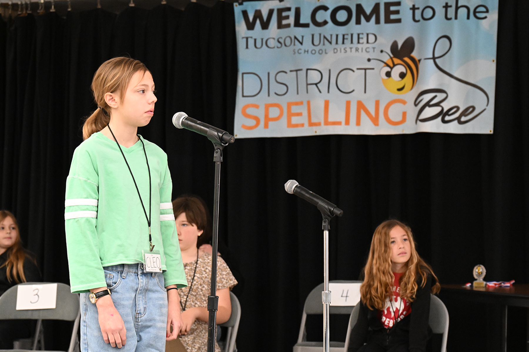 A girl in a green shirt stands at the microphone, preparing to spell her next word