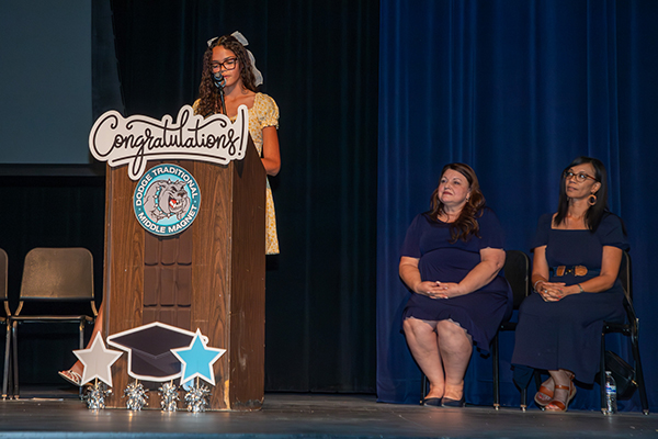 A teen girl addresses her class at the podium, while two women sit behind her