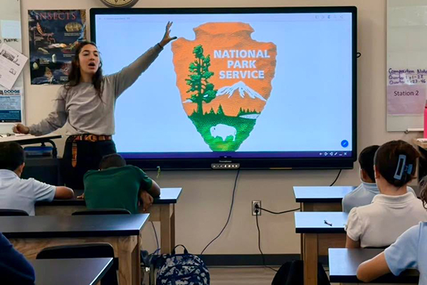 A woman holds her arm up while talking to students in front of the National Park Service logo on a smartboard